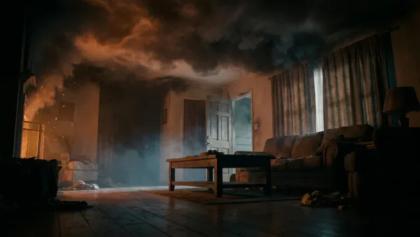 A dark living room early in a house fire, seen from floor level: a thick black smoke layer presses down from the ceiling and glows orange, while a thin band of clearer air remains near the floor over a sofa, coffee table, and doorway — showing why smoke inhalation strikes before the flames reach you.
